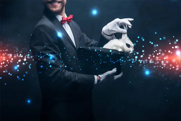 Abgeschnittene Ansicht eines lächelnden Zauberers, der einen Trick mit weißem Kaninchen im Hut zeigt, in einem dunklen Raum mit Rauch und glühender Illustration — Stockfoto