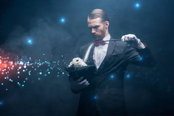 Serious magician in suit showing trick with wand and white rabbit in hat, dark room with smoke and glowing illustration — Stock Photo