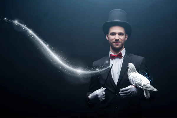 Happy magician in hat making abracadabra with dove and wand in dark room with smoke and glowing illustration — Stock Photo
