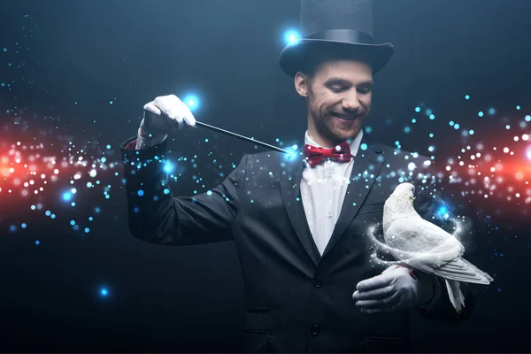 Happy magician in hat making abracadabra with dove and wand in dark room with smoke and glowing illustration — Stock Photo