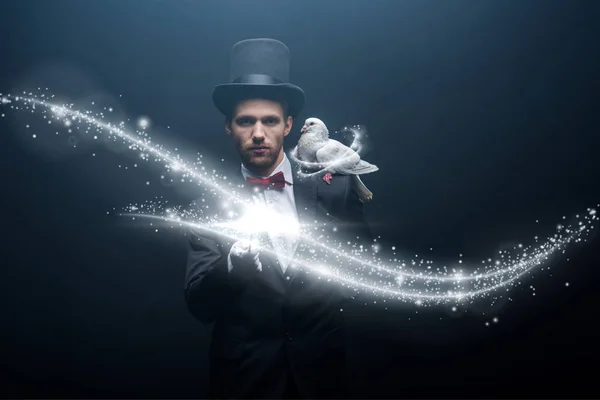 Dove sitting on shoulder of young magician in hat with wand in dark room with smoke and glowing illustration — Stock Photo