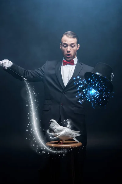 Shocked magician showing trick with dove, wand and hat in dark room with smoke and glowing illustration — Stock Photo