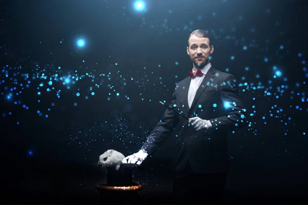 Happy magician in suit showing trick with wand and white rabbit in hat, dark room with smoke and glowing illustration — Stock Photo