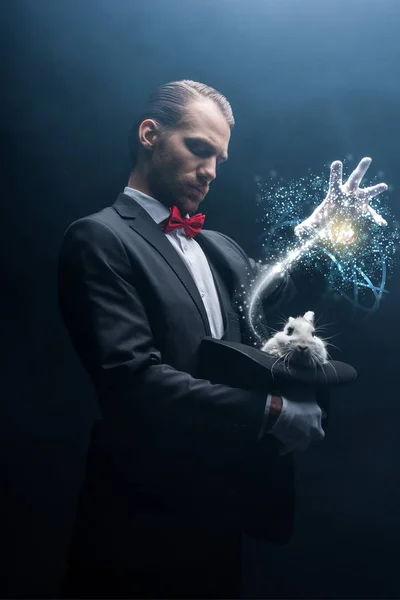 Профессиональный фокусник жестом над белым кроликом в шляпе, темная комната с дымом и светящиеся иллюстрации — стоковое фото