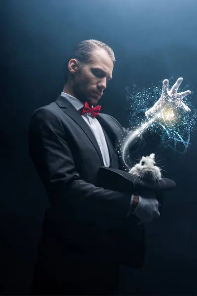 Professional magician gesturing over white rabbit in hat, dark room with smoke and glowing illustration — Stock Photo