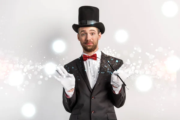 Magicien adulte avec geste hausseur d'épaules tenant baguette sur gris avec illustration lumineuse — Photo de stock