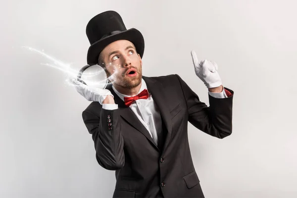 Professional magician pointing up and listening to magic ball isolated on grey with glowing illustration — Stock Photo