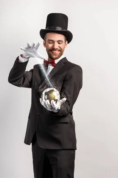 Happy professional magician holding magic ball isolated on grey with glowing illustration — Stock Photo