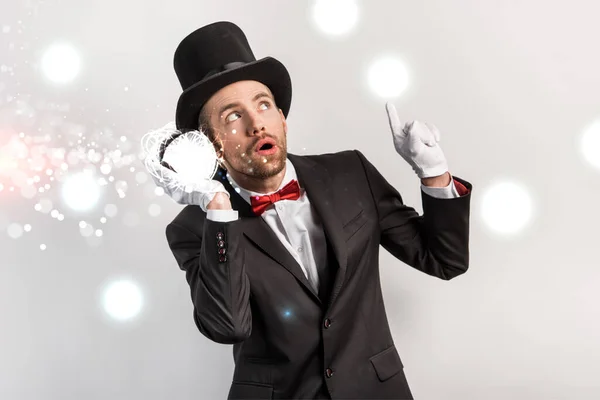 Professional magician pointing up and listening to magic ball isolated on grey with glowing illustration — Stock Photo
