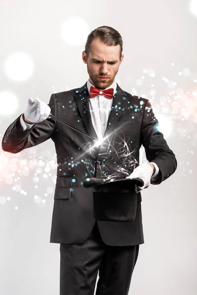 Confused magician in suit making trick hat and wand isolated on grey with glowing illustration — Stock Photo