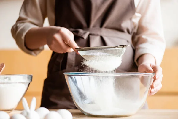 Geschnittene Ansicht einer Frau, die Mehl in Glasschüssel in der Nähe von Eiern und dekorativem Kaninchen siebte — Stockfoto