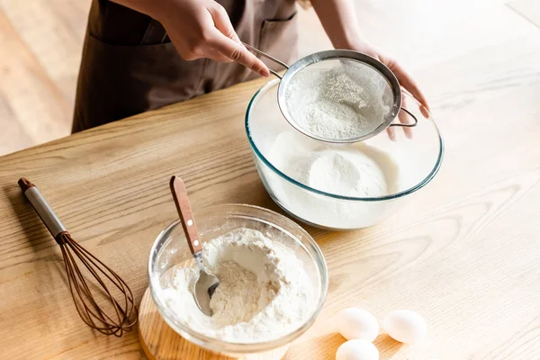 Vista ritagliata di giovane donna setacciare farina in ciotola di vetro vicino frusta, cucchiaio e uova — Foto stock