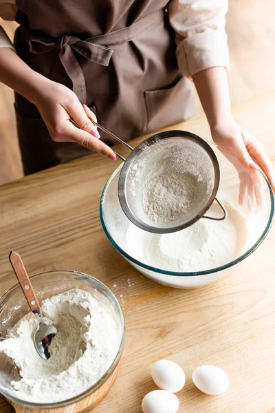 Top view of young woman sieving flour in glass bowl near raw eggs — Stock Photo