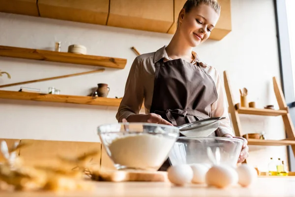 Selektiver Fokus einer glücklichen Frau, die Mehl in Glasschüssel neben rohen Eiern auf dem Tisch siebte — Stockfoto