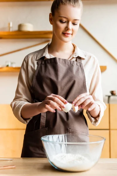 Attraktive Frau in Schürze, die Ei in Schüssel mit Mehl hinzufügt — Stockfoto