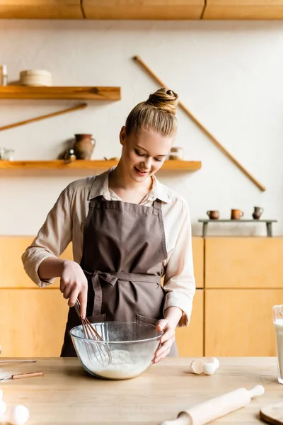 Happy woman in apron holding whisk near bowl with flour — Stock Photo