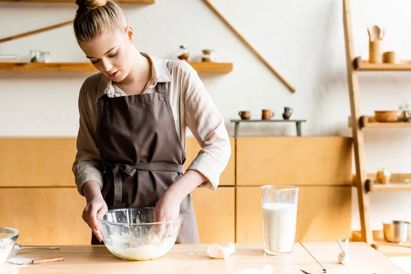 Attraktive Frau in Schürze knetet Teig mit der Hand in der Nähe Krug mit Milch und Osterhasenfigur — Stockfoto