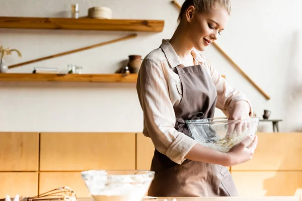 Happy woman in apron holding bowl while kneading dough — Stock Photo
