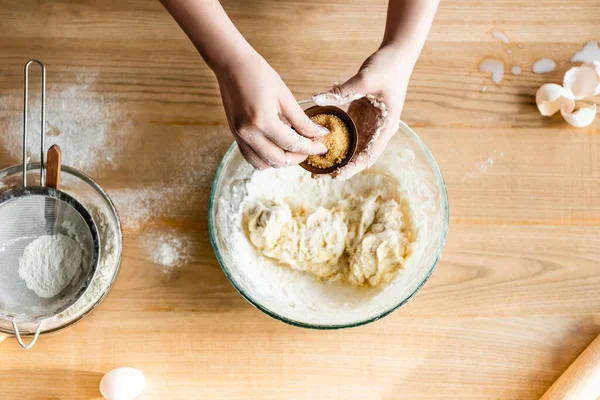 Top view of woman holding bowl with brown sugar above dough in bowl, raw eggs and flour — Stock Photo