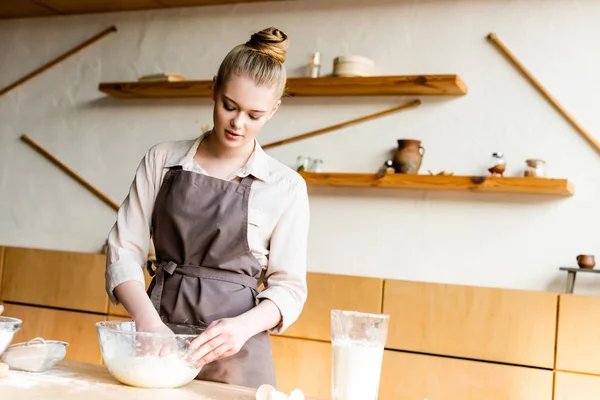 Atractiva mujer amasando masa en un tazón cerca de la jarra con leche - foto de stock