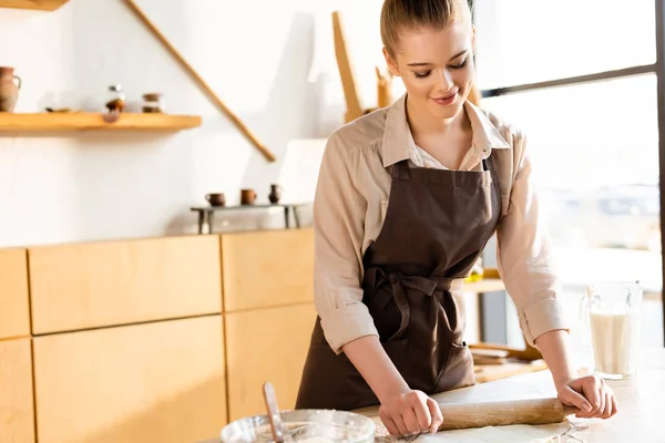 Fille heureuse dans tablier déroulant la pâte près de cruche avec du lait — Photo de stock