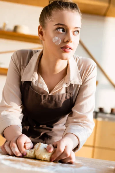 Beautiful young woman in apron kneading dough and looking away — Stock Photo