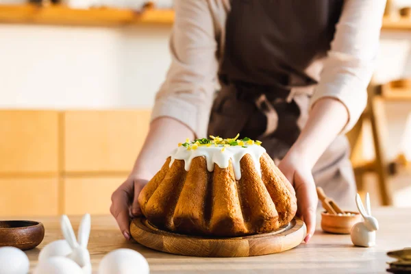 Vista recortada de la mujer sosteniendo pastel de Pascua cerca de figurillas con conejos decorativos - foto de stock