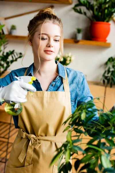 Beautiful woman in apron holding spray bottle with water near green leaves — Stock Photo