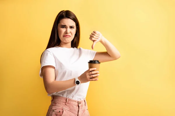 Menina descontente mostrando polegar para baixo e sorridente enquanto segurando café para ir isolado no amarelo — Fotografia de Stock