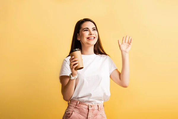 Menina feliz segurando café para ir e acenando a mão enquanto olha para longe isolado no amarelo — Fotografia de Stock