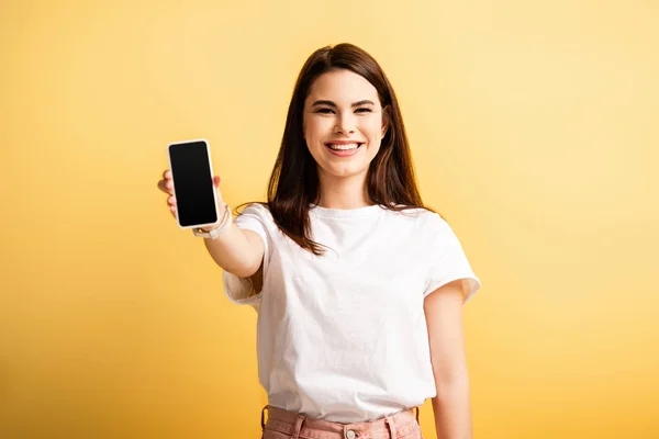 Chica feliz mostrando teléfono inteligente con pantalla en blanco mientras sonríe a la cámara en el fondo amarillo - foto de stock