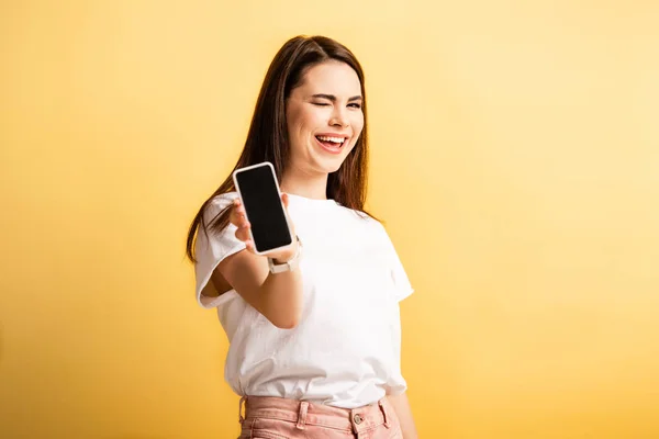 Menina alegre piscando para a câmera ao mostrar smartphone com tela em branco isolado no amarelo — Fotografia de Stock