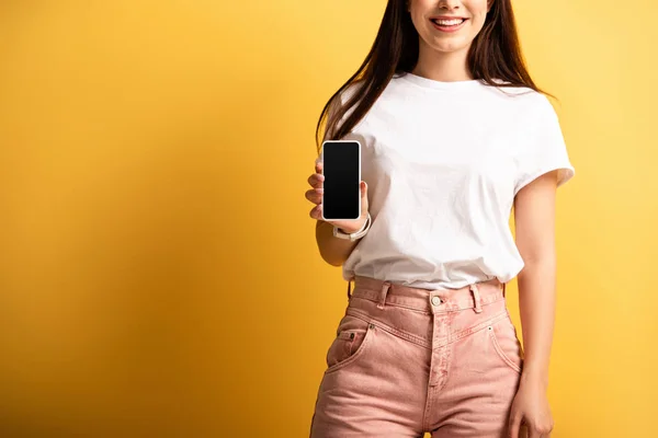 Vista recortada de la chica sonriente que muestra el teléfono inteligente con pantalla en blanco sobre fondo amarillo - foto de stock