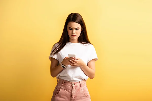 Fille mécontente fronçant les sourcils tandis que la messagerie sur smartphone isolé sur jaune — Photo de stock