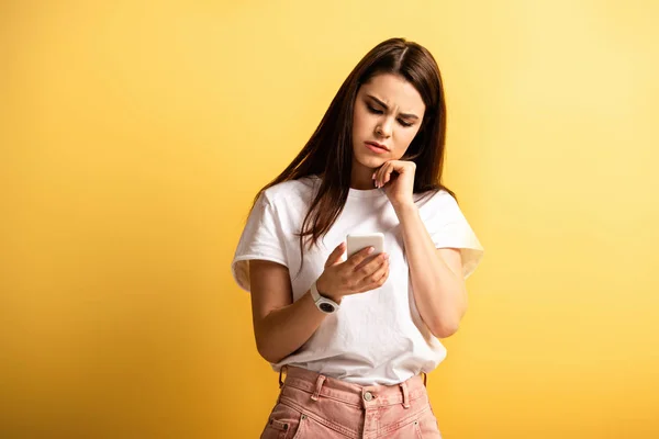 Chica reflexiva sosteniendo la mano cerca de la barbilla mientras chatea en el teléfono inteligente sobre fondo amarillo - foto de stock