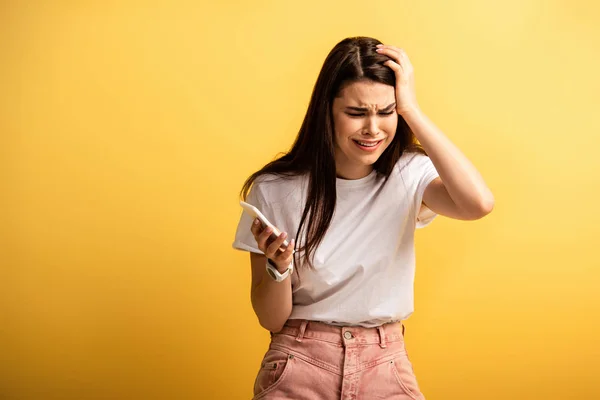 Upset girl touching head while holding smartphone on yellow background — Stock Photo