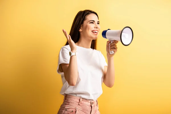 Happy girl speaking in megaphone while standing with open arm and looking away on yellow background — Stock Photo