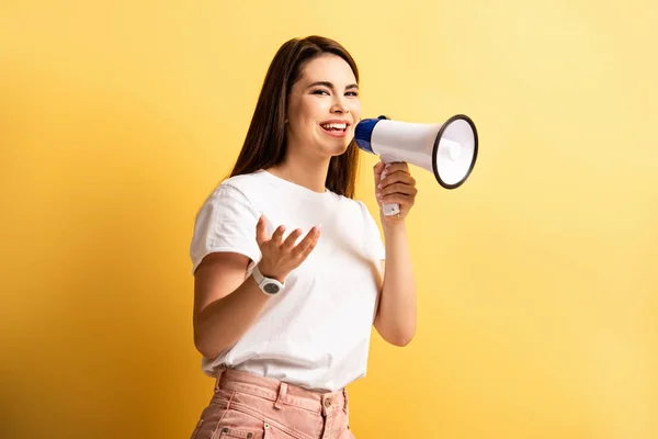 Cheerful girl speaking in megaphone while standing with open arm on yellow background — Stock Photo