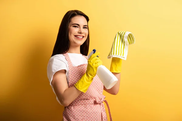 Heureuse femme au foyer en tablier et gants en caoutchouc tenant bouteille de pulvérisation et chiffon sur fond jaune — Photo de stock