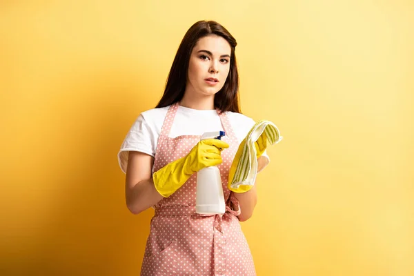 Ama de casa pensativa en delantal y guantes de goma rociando detergente sobre trapo sobre fondo amarillo - foto de stock