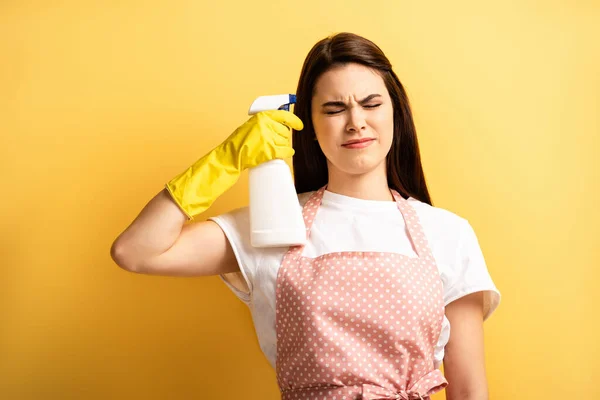 Jovem dona de casa em avental e luvas de borracha imitando auto-matança com frasco de spray com olhos fechados no fundo amarelo — Fotografia de Stock