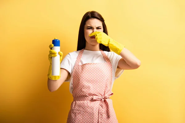 Displeased housewife in apron and rubber gloves plugging hose with hand while holding air freshener on yellow background — Stock Photo
