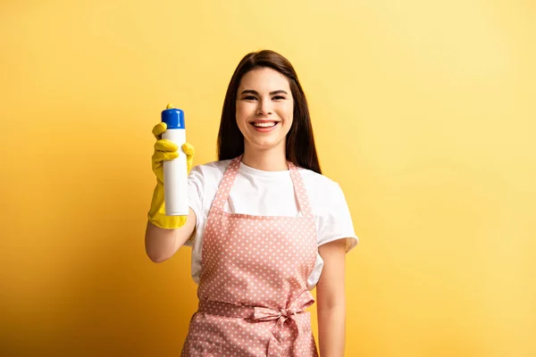 Heureuse femme au foyer dans tablier et gants en caoutchouc regardant la caméra tout en tenant désodorisant sur fond jaune — Photo de stock