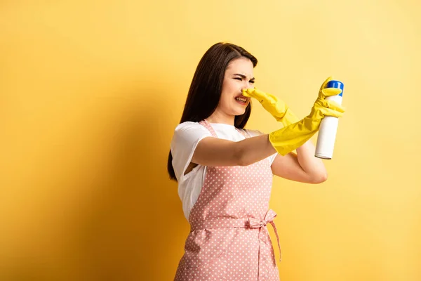 Dona de casa descontente em avental e luvas de borracha plugando mangueira com a mão enquanto pulverizando ambientador de ar no fundo amarelo — Fotografia de Stock