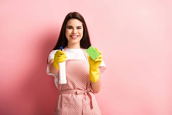 Happy housewife in apron and rubber gloves holding spray bottle and sponge on pink background — Stock Photo