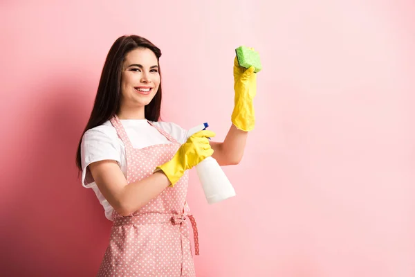 Весёлая домохозяйка в фартуке и резиновых перчатках, держащая бутылку с брызгами и губку на розовом фоне — стоковое фото