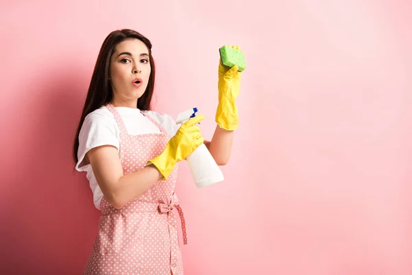 Dona de casa surpresa no avental e luvas de borracha segurando spray garrafa e esponja no fundo rosa — Fotografia de Stock