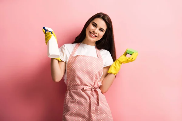 Happy housewife in apron and rubber gloves holding spray bottle and sponge on pink background — Stock Photo