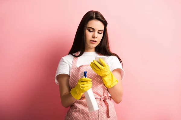 Ama de casa enfocada en delantal y guantes de goma charlando en el teléfono inteligente mientras sostiene la botella de spray sobre fondo rosa - foto de stock