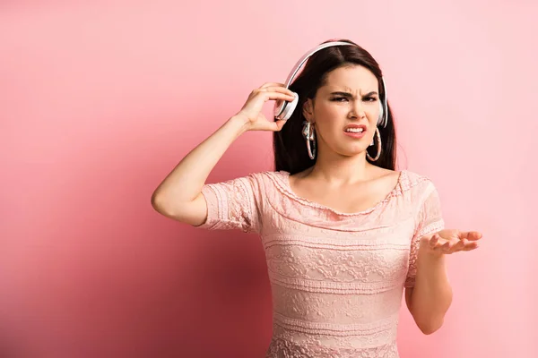 Chica disgustada tocando auriculares inalámbricos mientras está de pie con el brazo abierto sobre fondo rosa - foto de stock